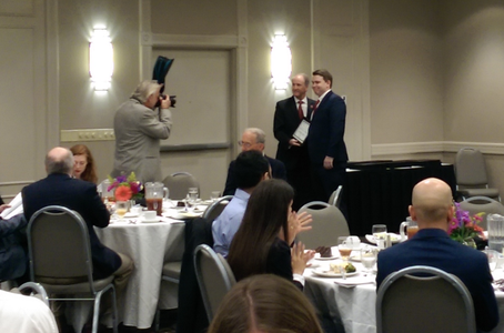 a man takes a photo of 2 people with an award in the middle of a dining area